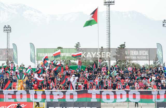 A stand filled with fans wearing red, white and green t-shirts and waving Palestinian flags.