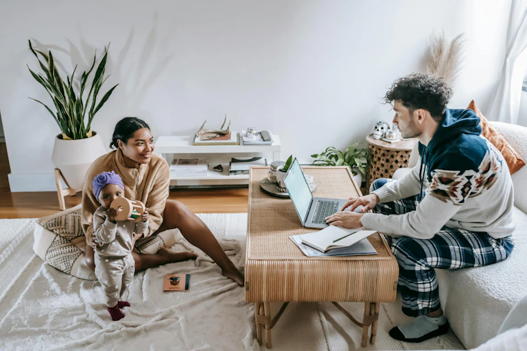 mother plays with child while her partner works on laptop nearby