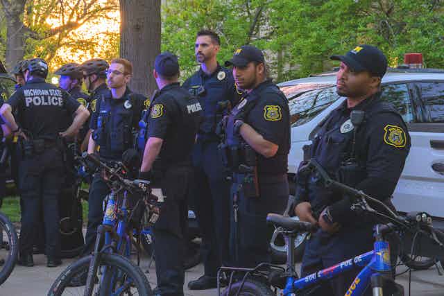 A small group of police officers, most of them white, gather near a police car and police bicycles.