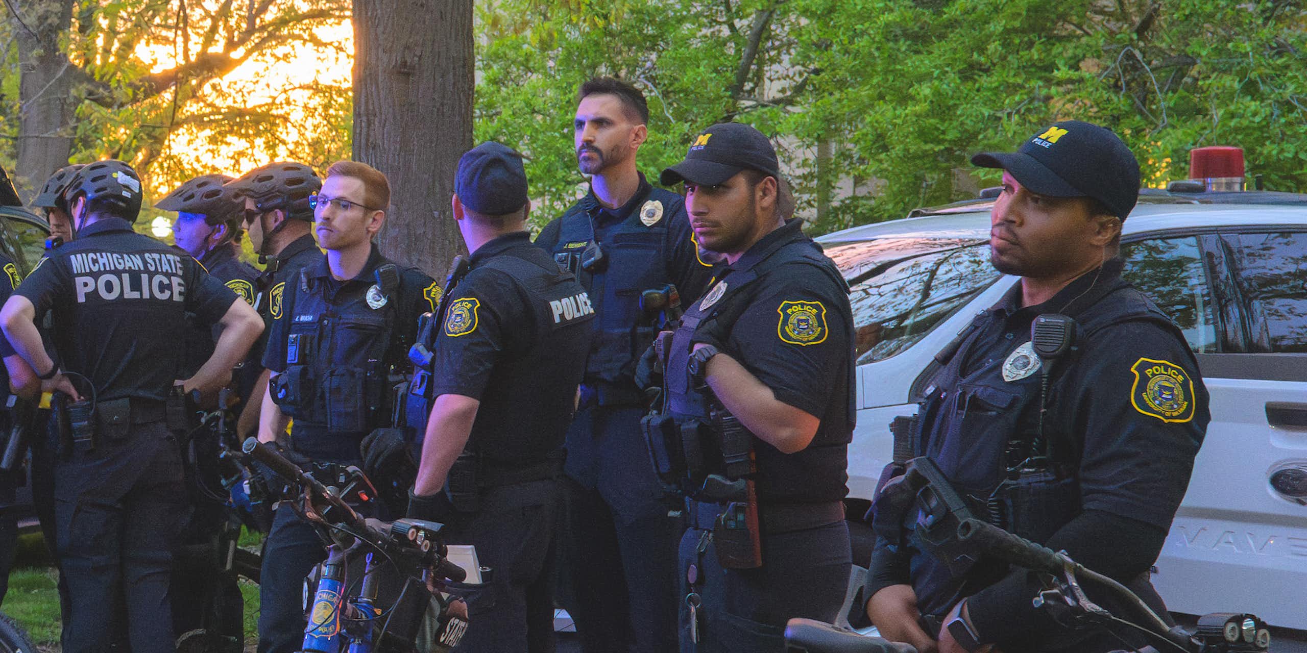 A small group of police officers, most of them white, gather near a police car and police bicycles.