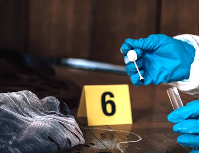 Photo of a pair of gloved hands taking a swab from a piece of fabric on a floor with a chalk outline and a numbered label.