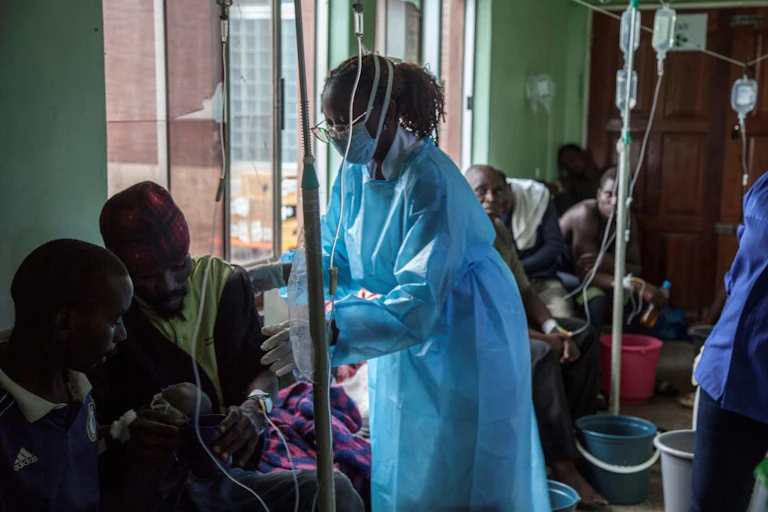 A woman in a blue medical gown and mask among people sitting in an area where medical drips are hanging from a line and buckets are on the floor