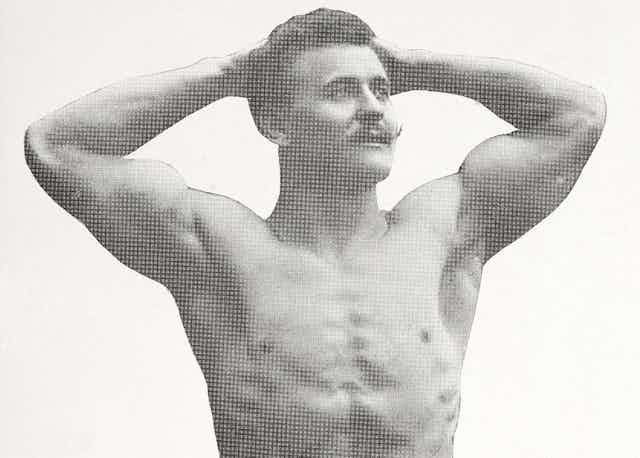 A black and white image of a man with a moustache, shirtless and muscular, his hands behind his head.