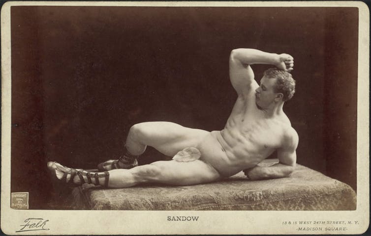 Un hombre musculoso descansa sobre un brazo, desnudo salvo por una hoja de parra en una fotografía antigua.