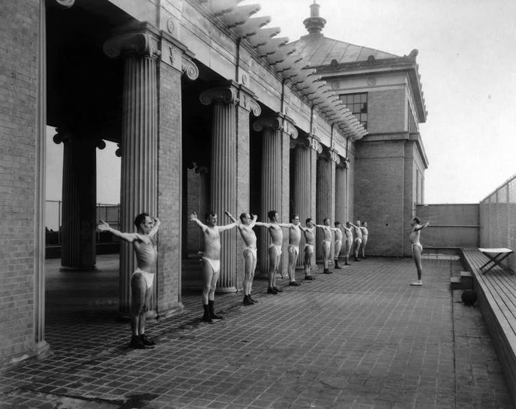 Black and white photo of a line of very thin men dressed in white underwear doing exercises on a formal patio featuring iconic columns.