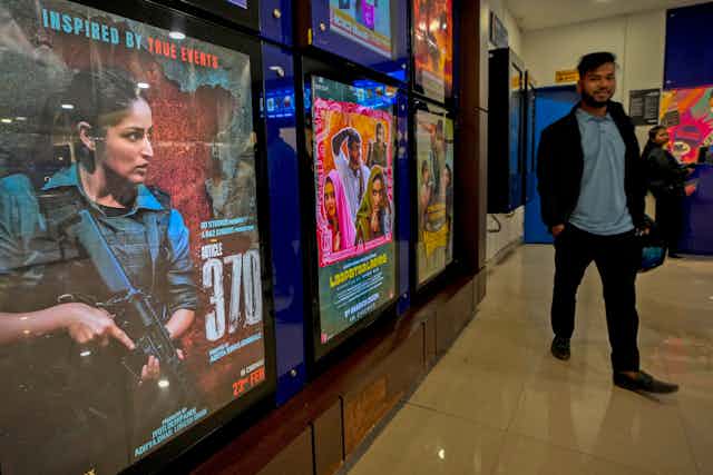 A movie poster shows a woman holding an automatic rifle. A man walks beside the film poster.