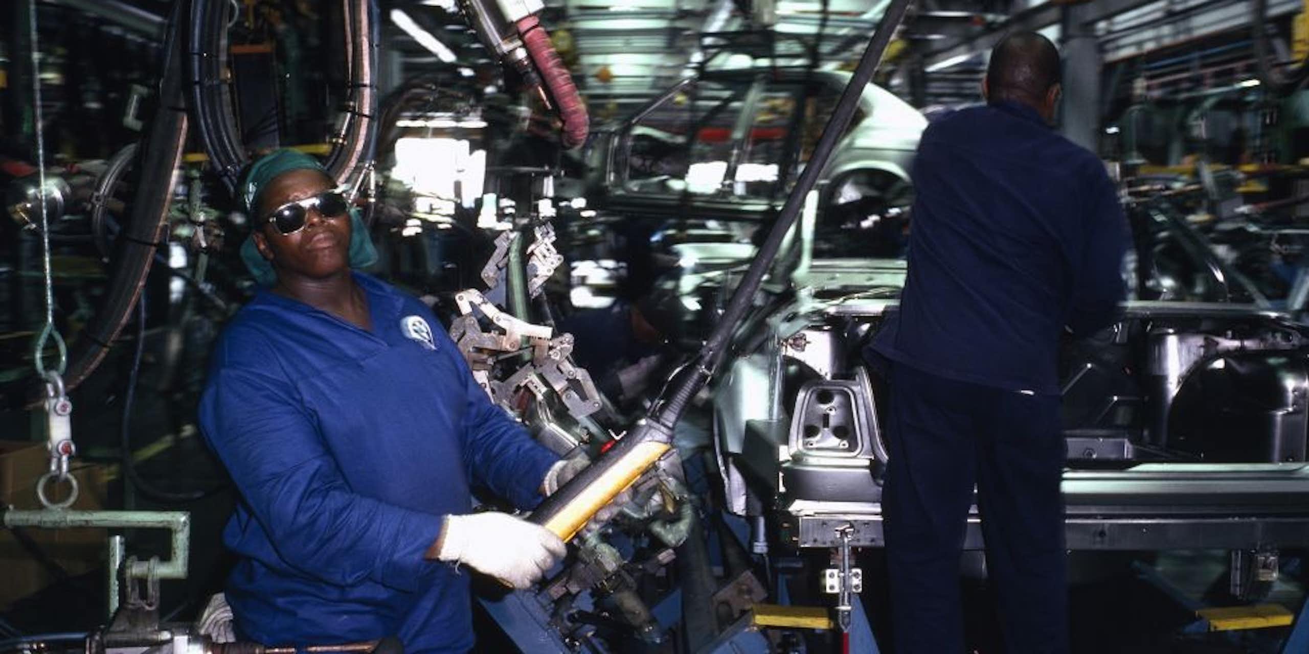 Men in blue overalls and protective glasses and gloves, amid machinery and the semi-built body of a vehicle