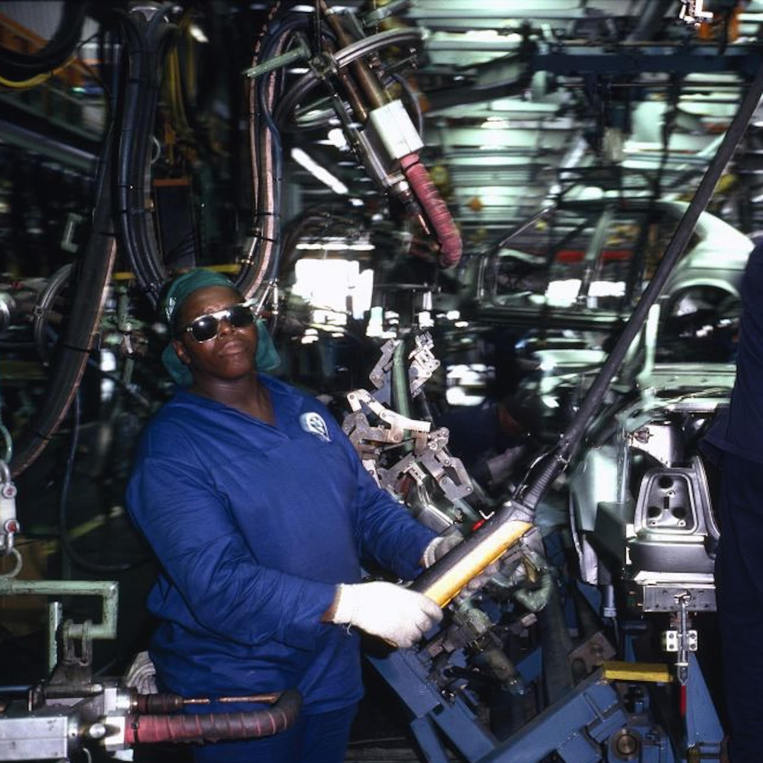 Men in blue overalls and protective glasses and gloves, amid machinery and the semi-built body of a vehicle