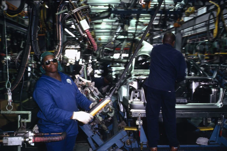 Men in blue overalls and protective glasses and gloves, amid machinery and the semi-built body of a vehicle