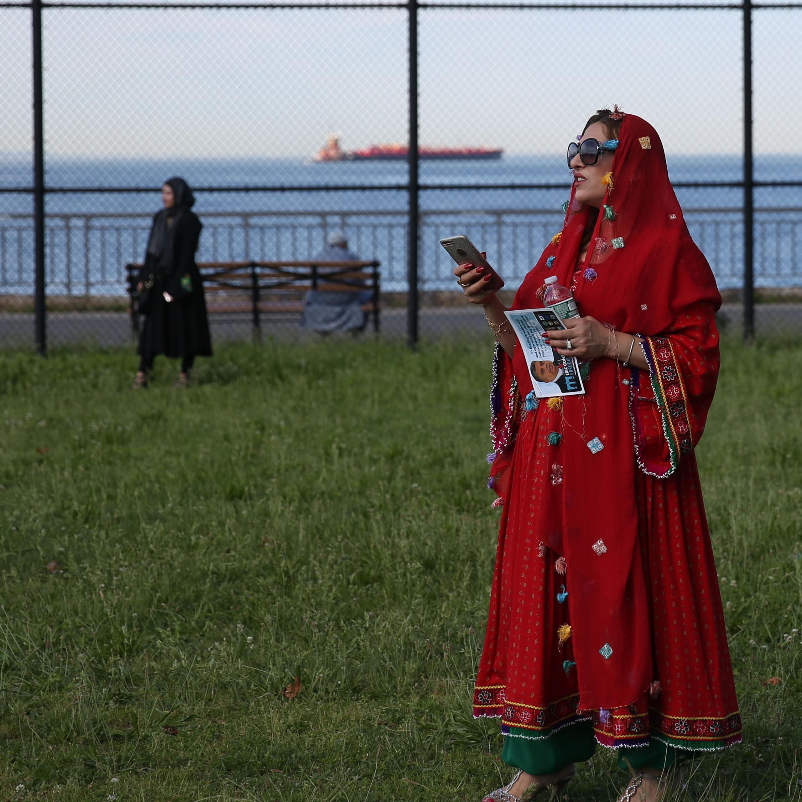 Muslim woman stands in a park holding a cel phone and looking off into the distance.