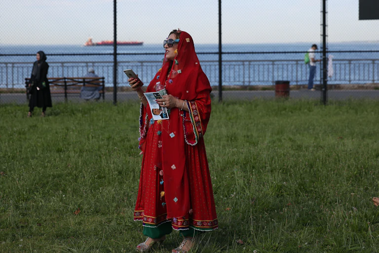 Muslim woman stands in a park holding a cel phone and looking off into the distance.