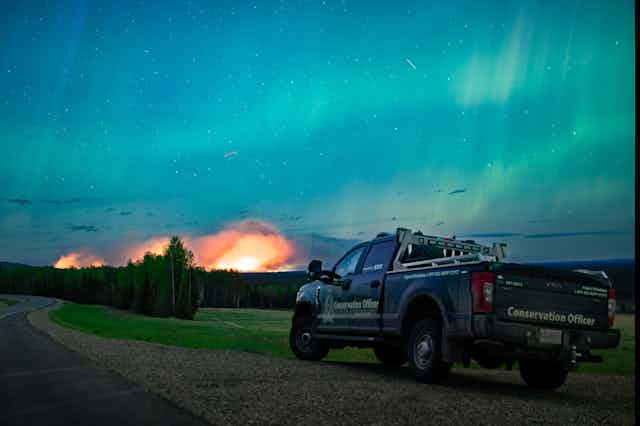 A truck is parked with a fire in the distance under an aurora filled night sky.