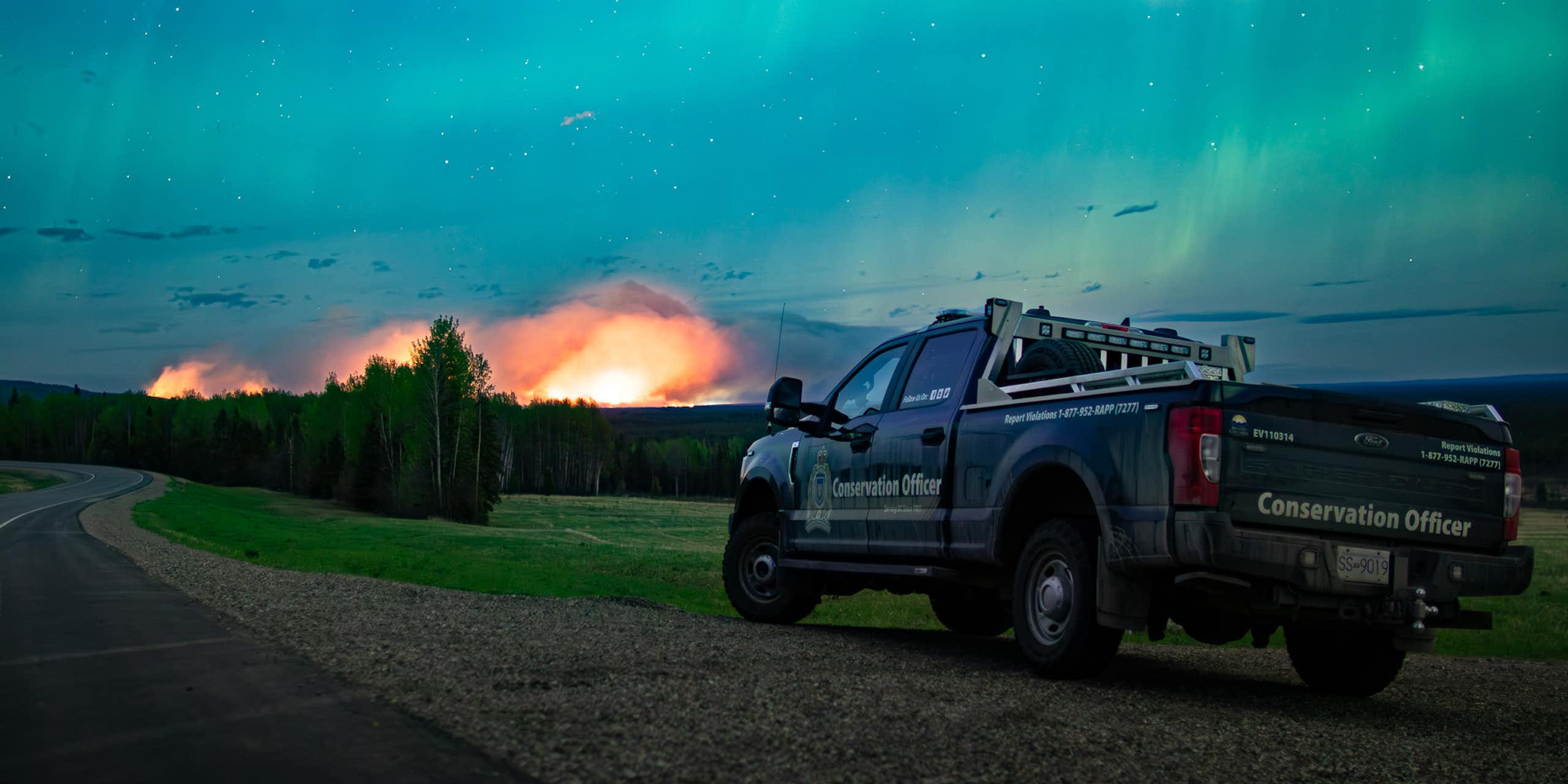 A truck is parked with a fire in the distance under an aurora filled night sky.