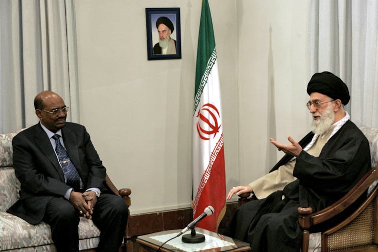 Two men sit in a room, one wearing a suit, the other wearing traditional Iranian clothing.