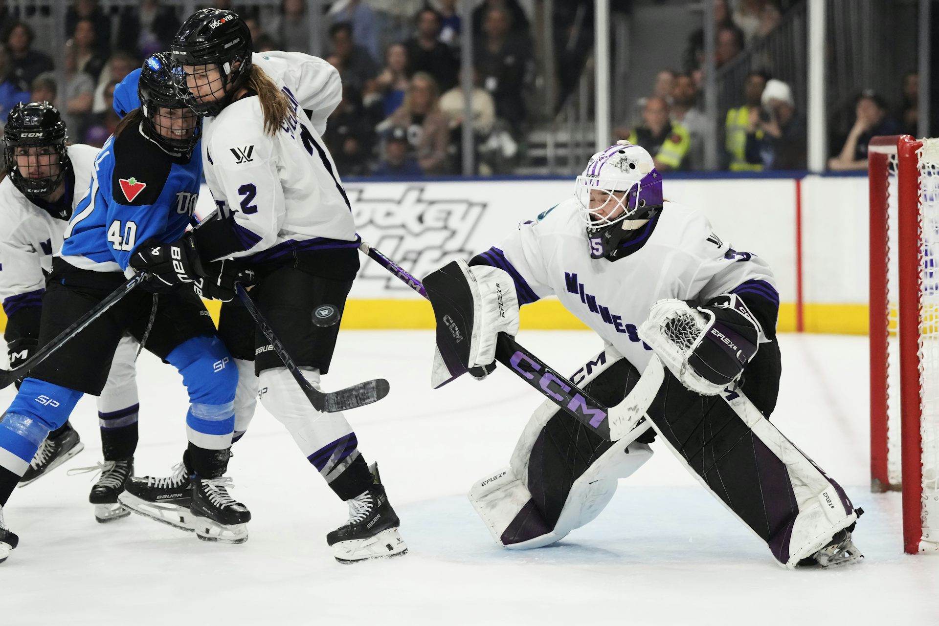 A hockey goalie crouches with their stick out as two other hockey players fight over a puck beside them