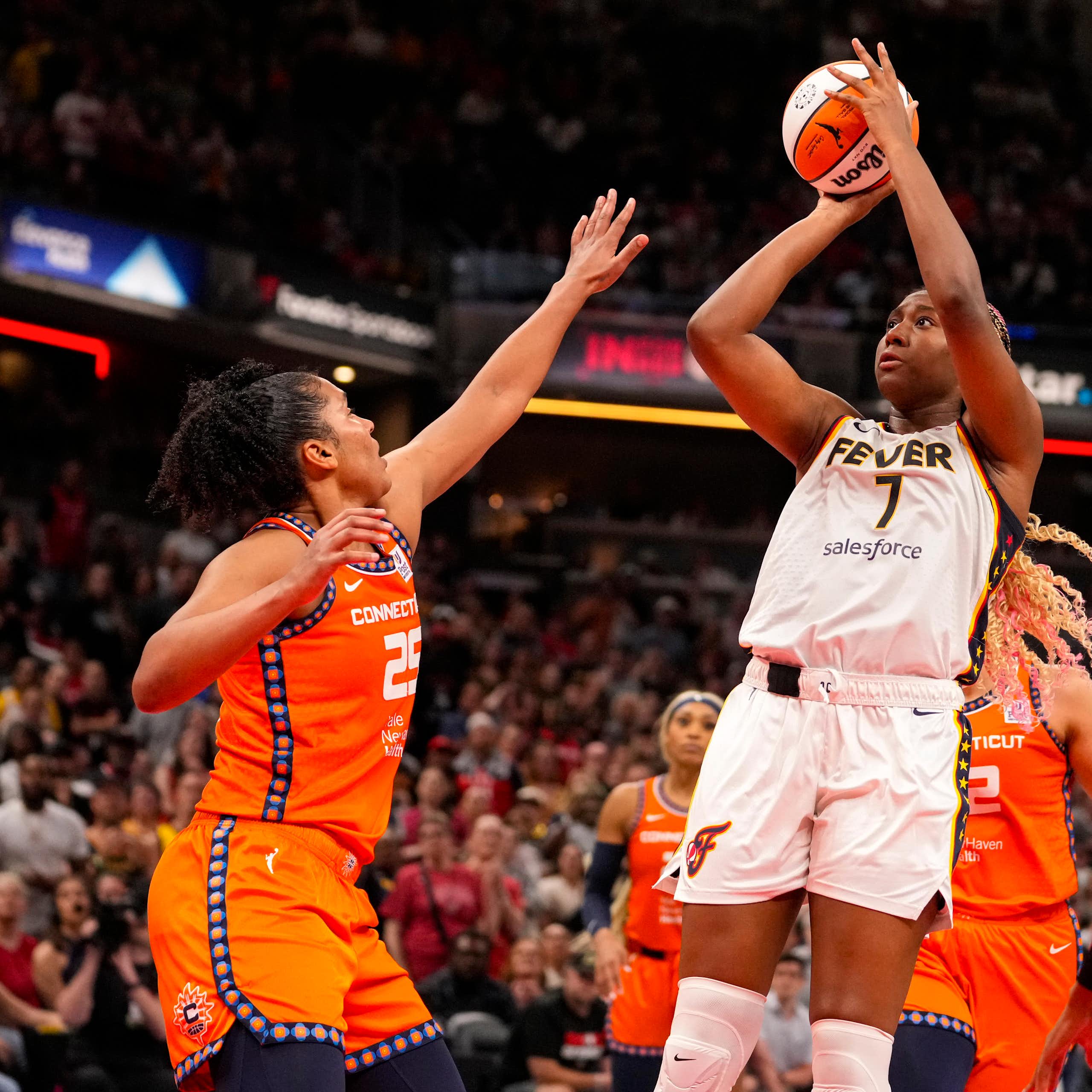 A Black woman in a basketball uniform jumps while doing a layup. Another woman reaches up to try and block the shot.