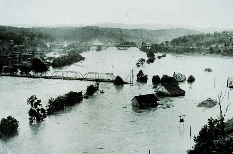 Wide river flooding surrounds industries and homes. Half a railroad span is gone.