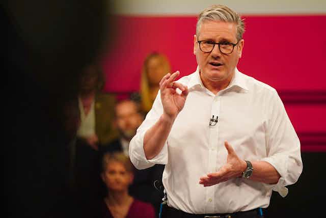 A man in a while shirt and black-rimmed glasses gesticulates as he talks