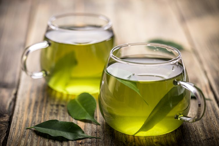 Two clear mugs of green tea, with leaves on wooden table