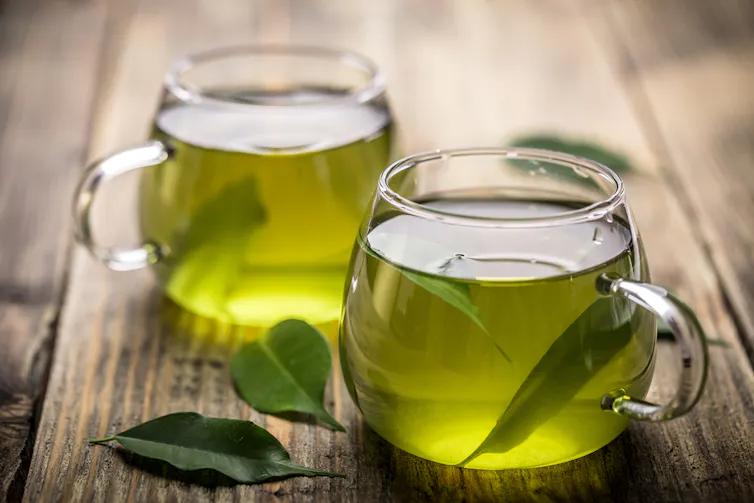 Two clear mugs of green tea, with leaves on wooden table