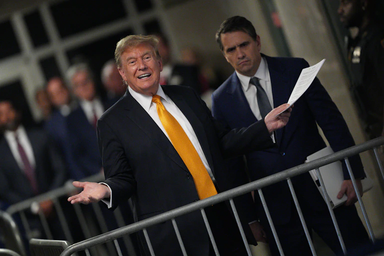 A middle-aged white man dressed in a business suit holds a sheet of paper as another man looks on.