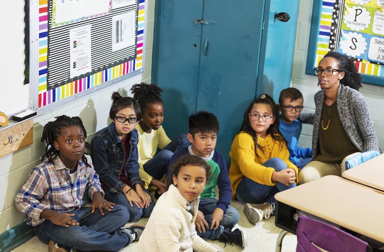 A Black woman wearing dark-rimmed glasses sits on the floor in the corner of a grade-school classroom next to a group of seven young, diverse children who also are seated. They are all looking in the same direction with serious expressions.