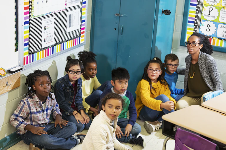 Students sit on the floor with their teacher looking worried.
