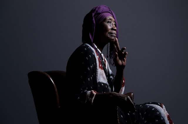Une femme africaine âgée est assise sur une chaise et pose pour un portrait de profil. Elle est vêtue d'une tenue africaine traditionnelle, porte la main à son menton et sourit légèrement.
