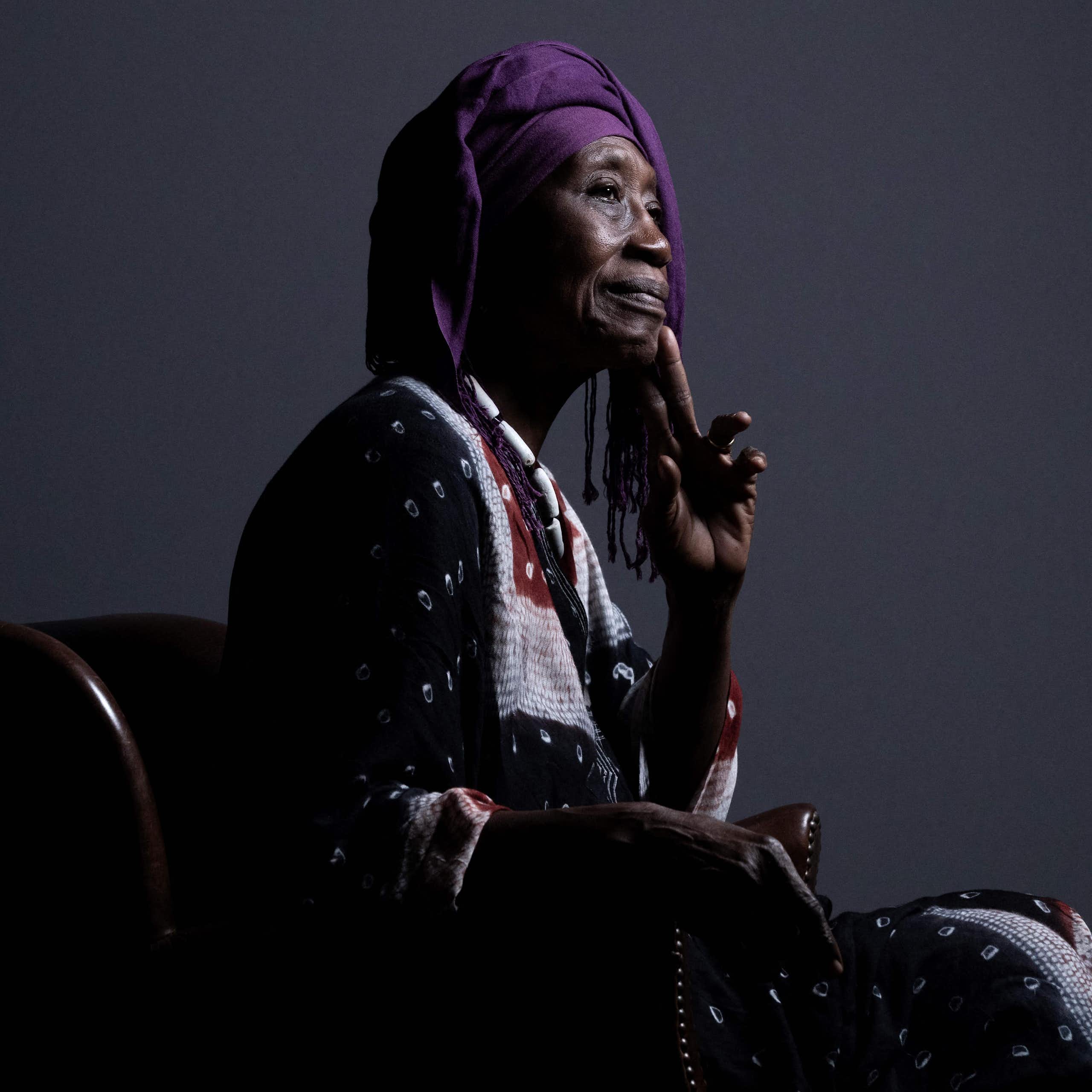 Une femme africaine âgée est assise sur une chaise et pose pour un portrait de profil. Elle est vêtue d'une tenue africaine traditionnelle, porte la main à son menton et sourit légèrement.