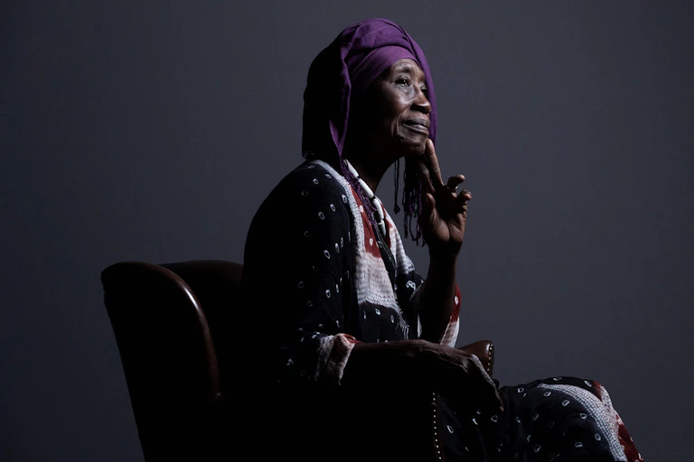 Une femme africaine âgée est assise sur une chaise et pose pour un portrait de profil. Elle est vêtue d'une tenue africaine traditionnelle, porte la main à son menton et sourit légèrement.