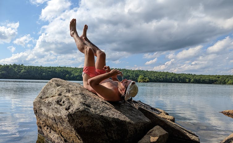 Man lying on a rock near a lake with his legs in the air and his posterior exposed to the sun.