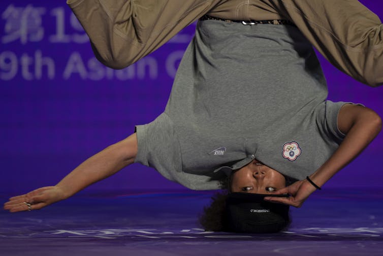 A breakdancer spins on his head, holding the brim of his hat with one arm and keeping the other outstretched.