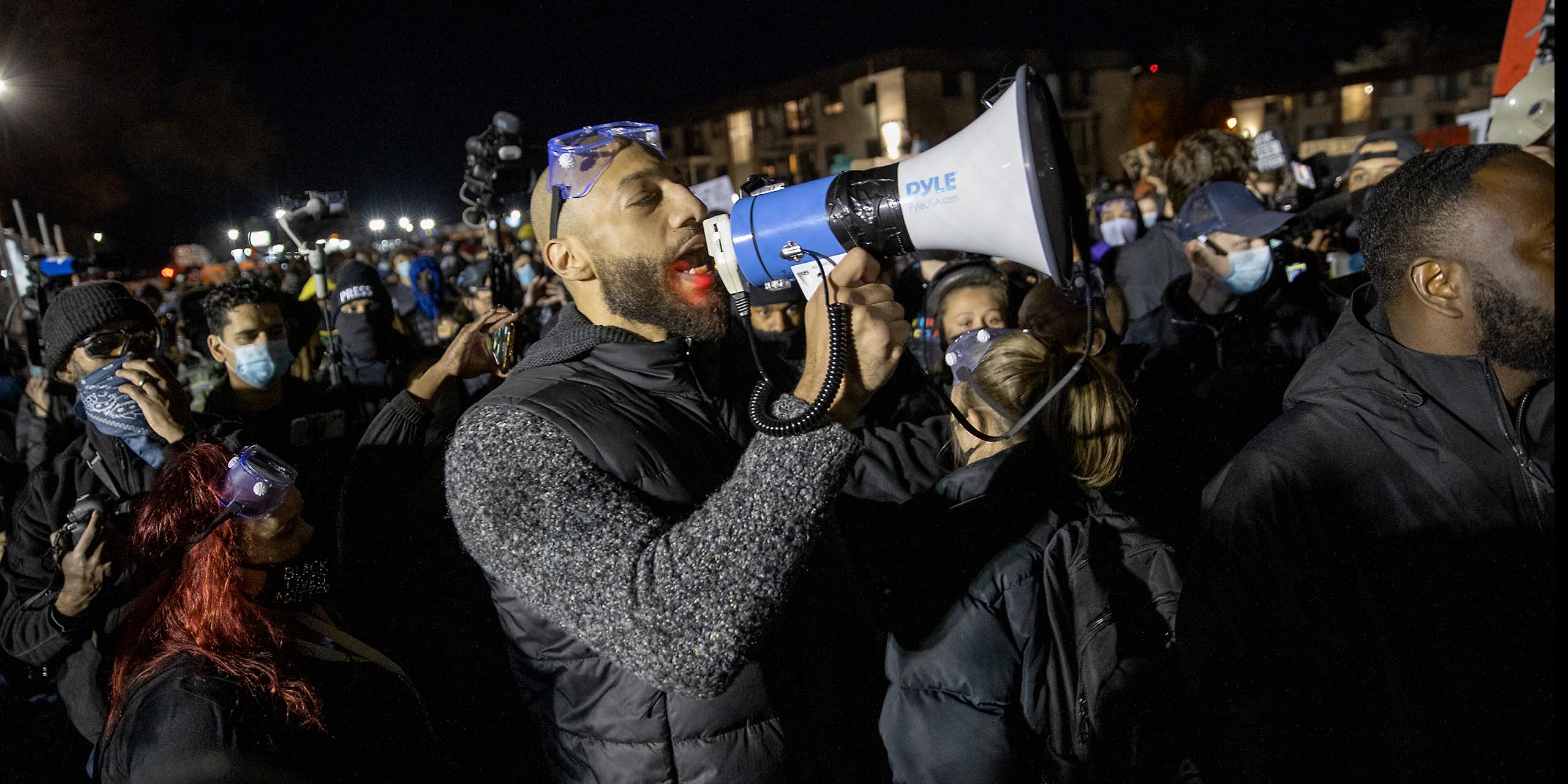 Bald black man yells into megaphone surrounded by a crowd.