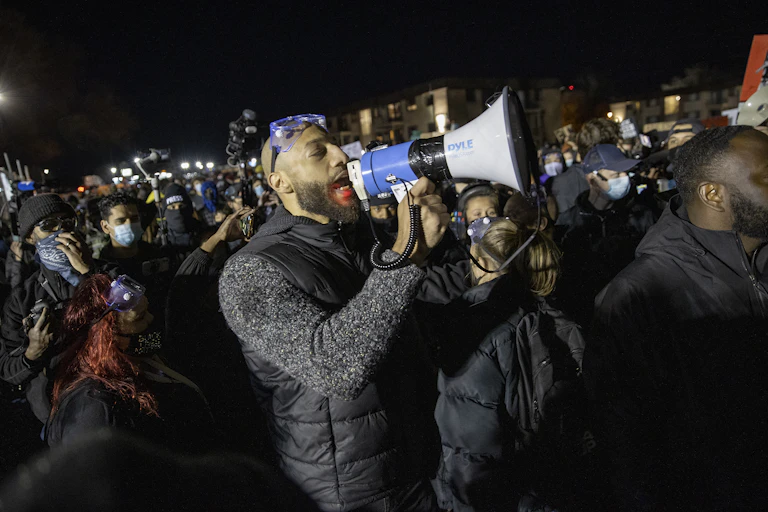Bald black man yells into megaphone surrounded by a crowd.