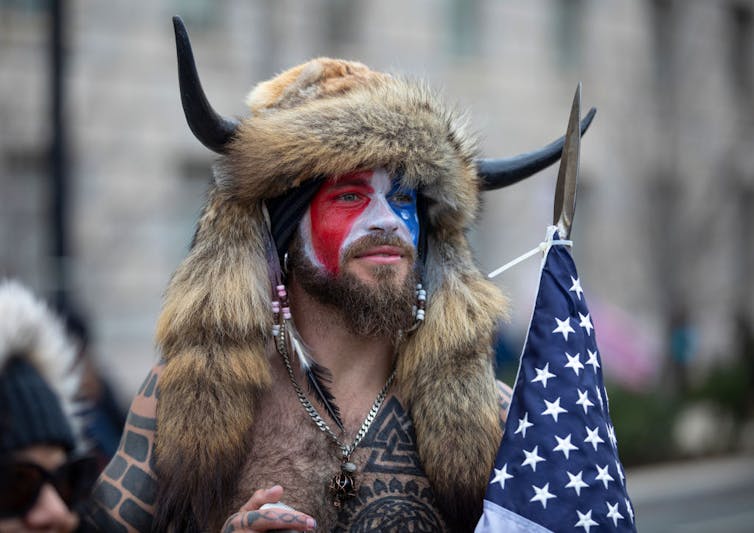 Hombre con la cara pintada con un sombrero de piel con cuernos y sosteniendo una bandera americana.