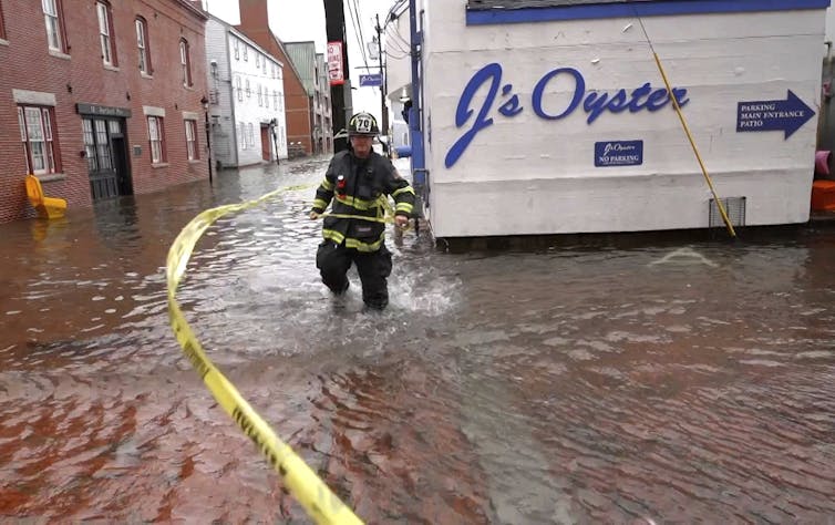 A firefighter walks through seawater running knee-deep in a commercial street with a sign for J's Oysters behind him.