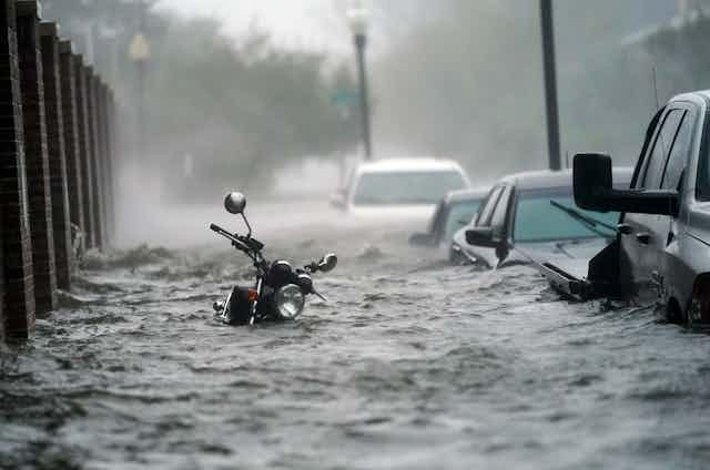 A flooded street during a hurricane with water up to the handle bars on a motorcycle and over the doors on cars parked along a street in Pensacola in 2020.