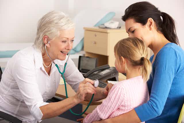Older woman GP using stethoscope on young girl sitting on mum's lap