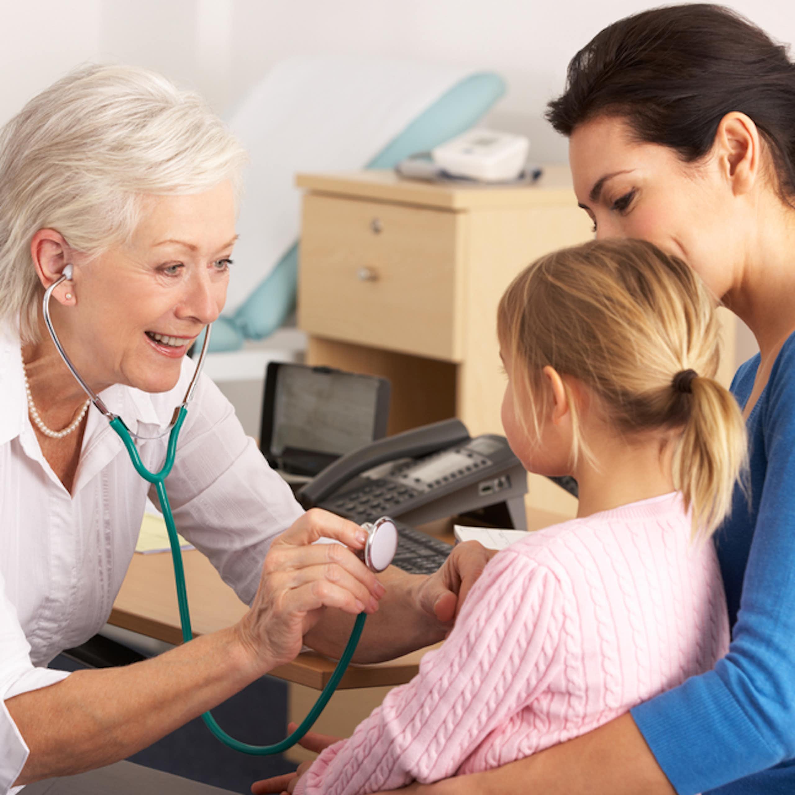 Older woman GP using stethoscope on young girl sitting on mum's lap