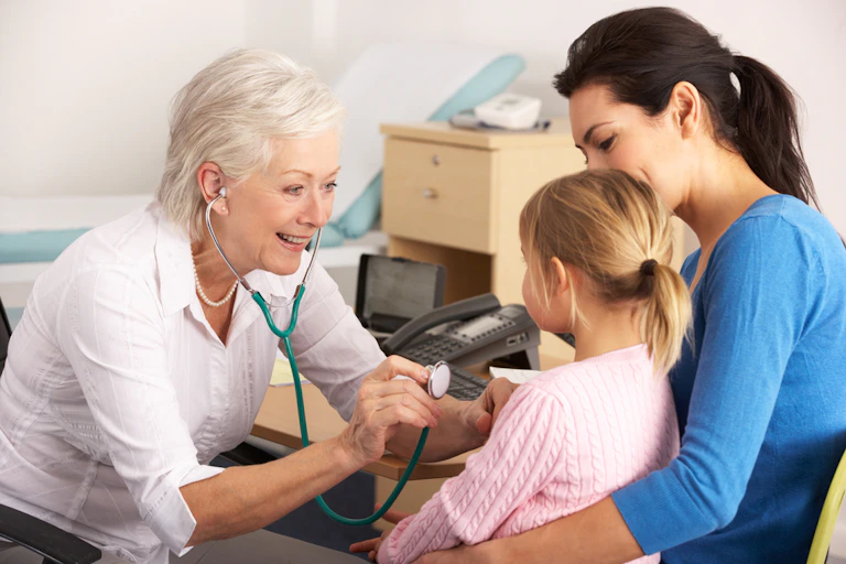 Older woman GP using stethoscope on young girl sitting on mum's lap