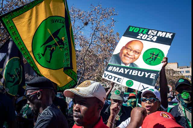 A small crowd of people in an urban setting hold up posters and banners - a poster reading "Vote MK 2024 #Mayibuye" and a green, gold and black banner with the letters "MK" below an illustration of a figure holding a spear.