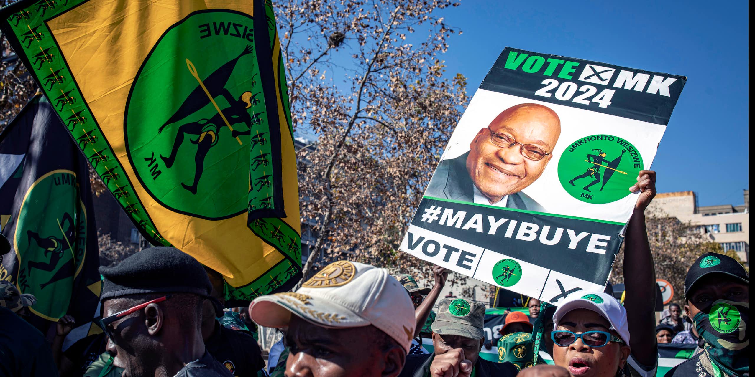 A small crowd of people in an urban setting hold up posters and banners - a poster reading "Vote MK 2024 #Mayibuye" and a green, gold and black banner with the letters "MK" below an illustration of a figure holding a spear.