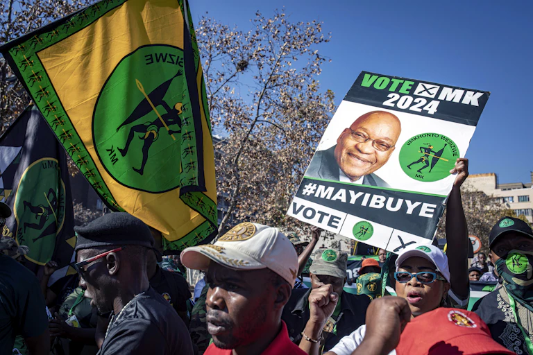 A small crowd of people in an urban setting hold up posters and banners - a poster reading "Vote MK 2024 #Mayibuye" and a green, gold and black banner with the letters "MK" below an illustration of a figure holding a spear.