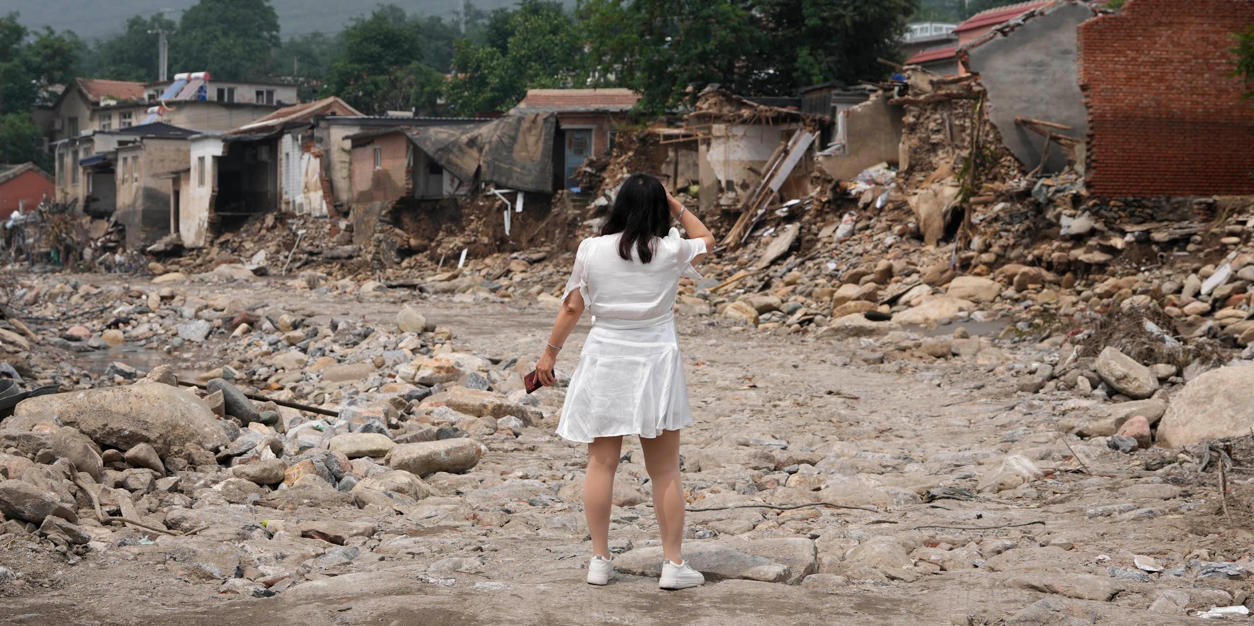 A woman in a white dress with her back to the camera stands in front of the wreckage of a row of houses