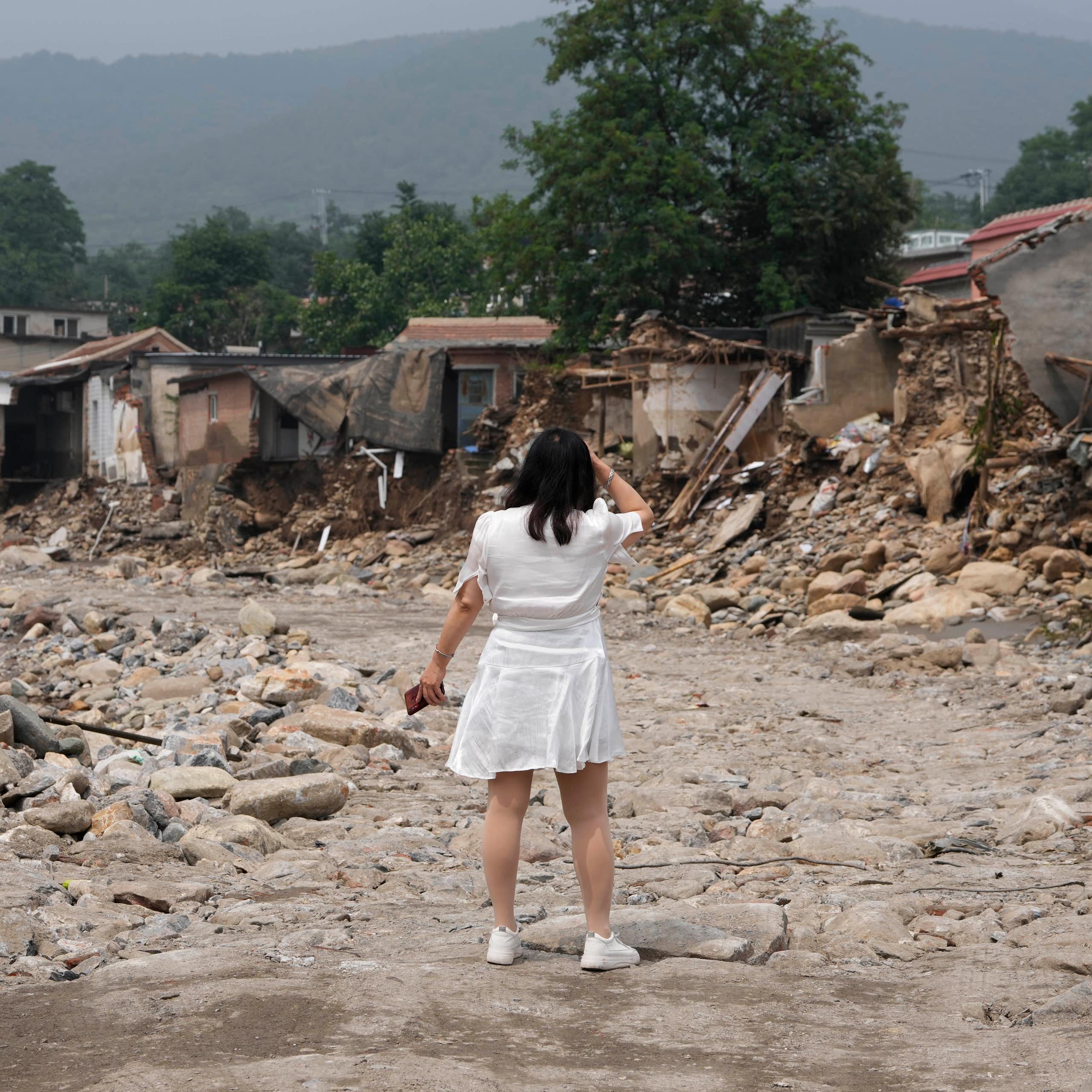 A woman in a white dress with her back to the camera stands in front of the wreckage of a row of houses