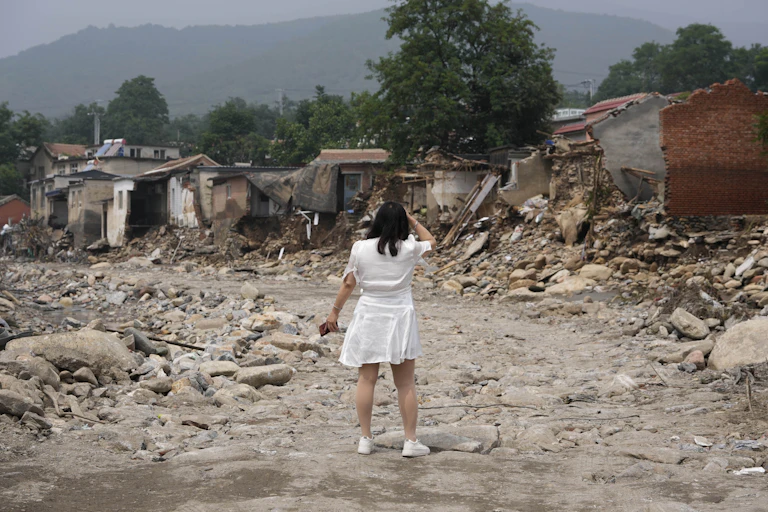 A woman in a white dress with her back to the camera stands in front of the wreckage of a row of houses