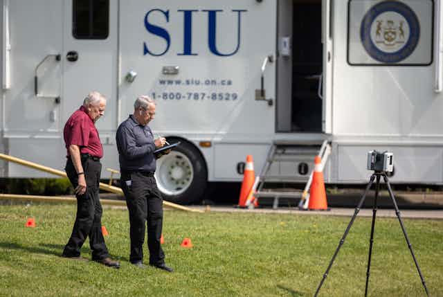Two older men stand on a grassy area. A truck with the letters SIU on it is behind them
