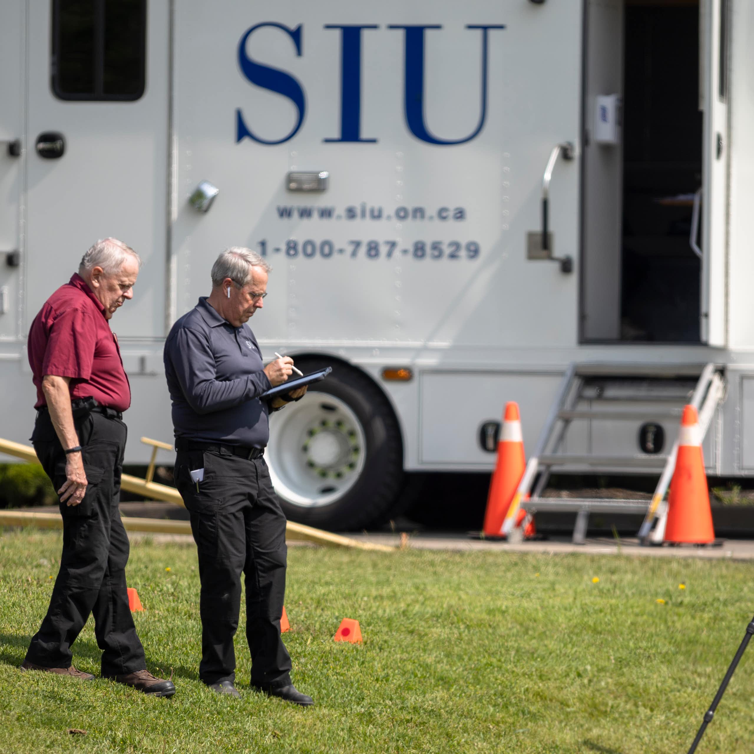 Two older men stand on a grassy area. A truck with the letters SIU on it is behind them