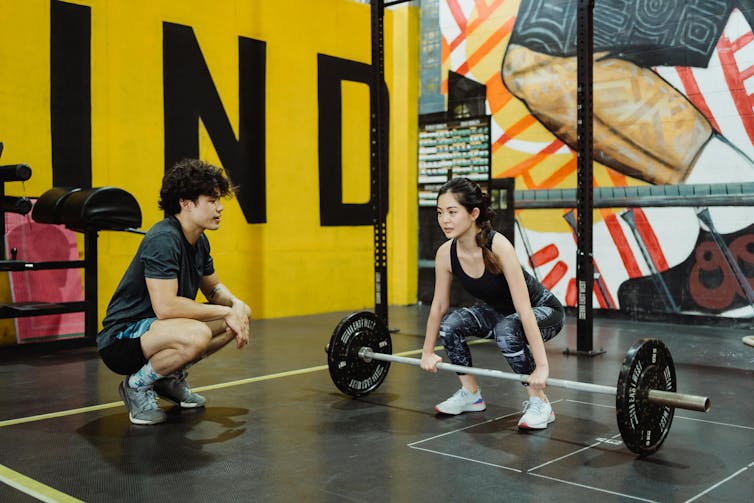 A woman sets up to lift a heavy weight while her trainer observes.