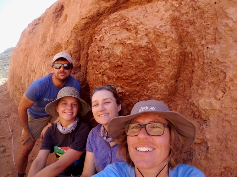Four people pose for a group selfie in front of a huge mound of red earth